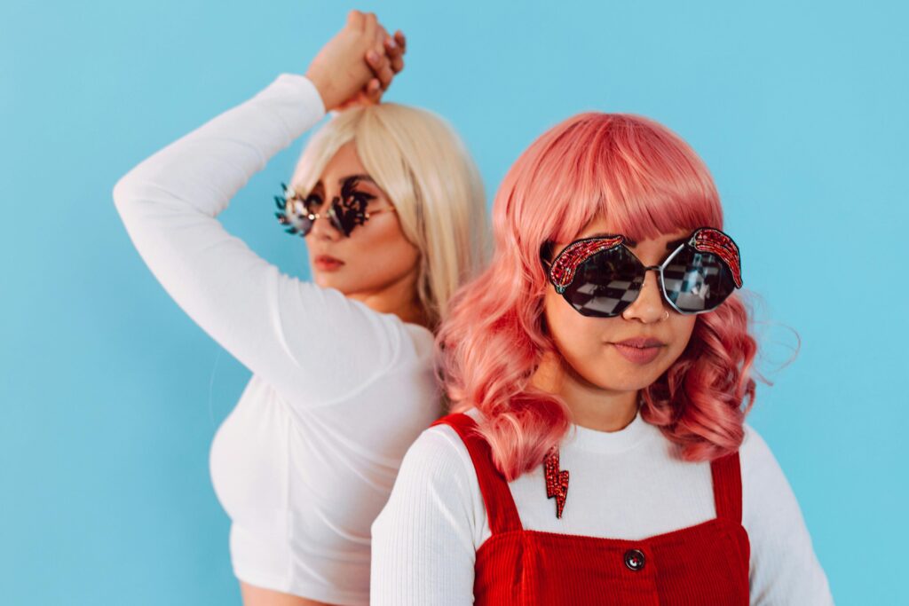 Two fashionable women with colorful hair and bold sunglasses against a blue background.