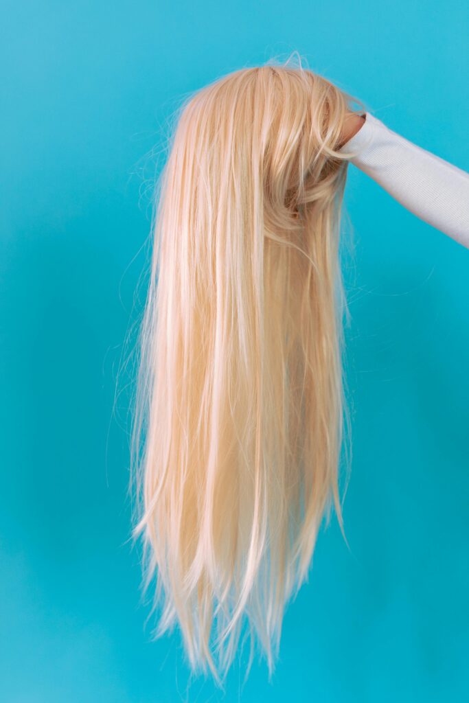 A blonde wig being held in a hand against a vivid blue background.