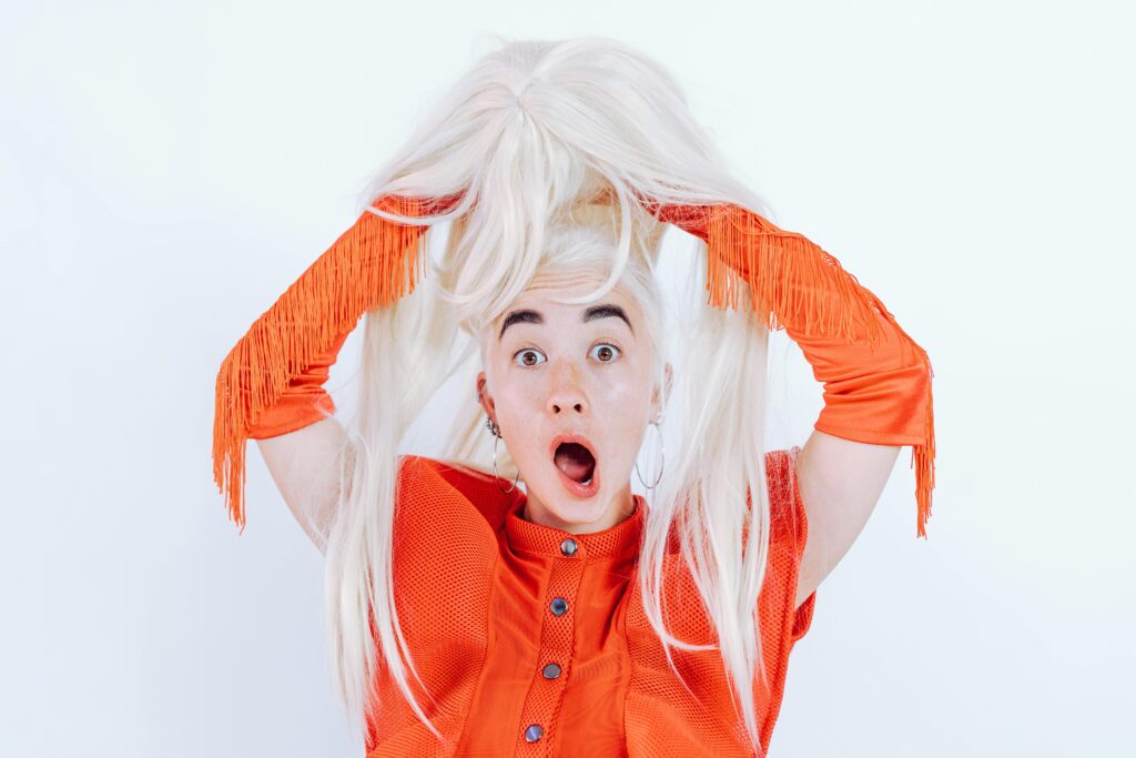 A startled woman in a vibrant orange outfit holding a blonde wig with a surprised expression.