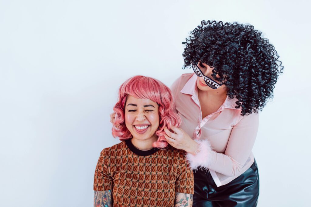 Two women enjoying a light-hearted moment, one wearing a pink wig and the other a black one, both laughing.
